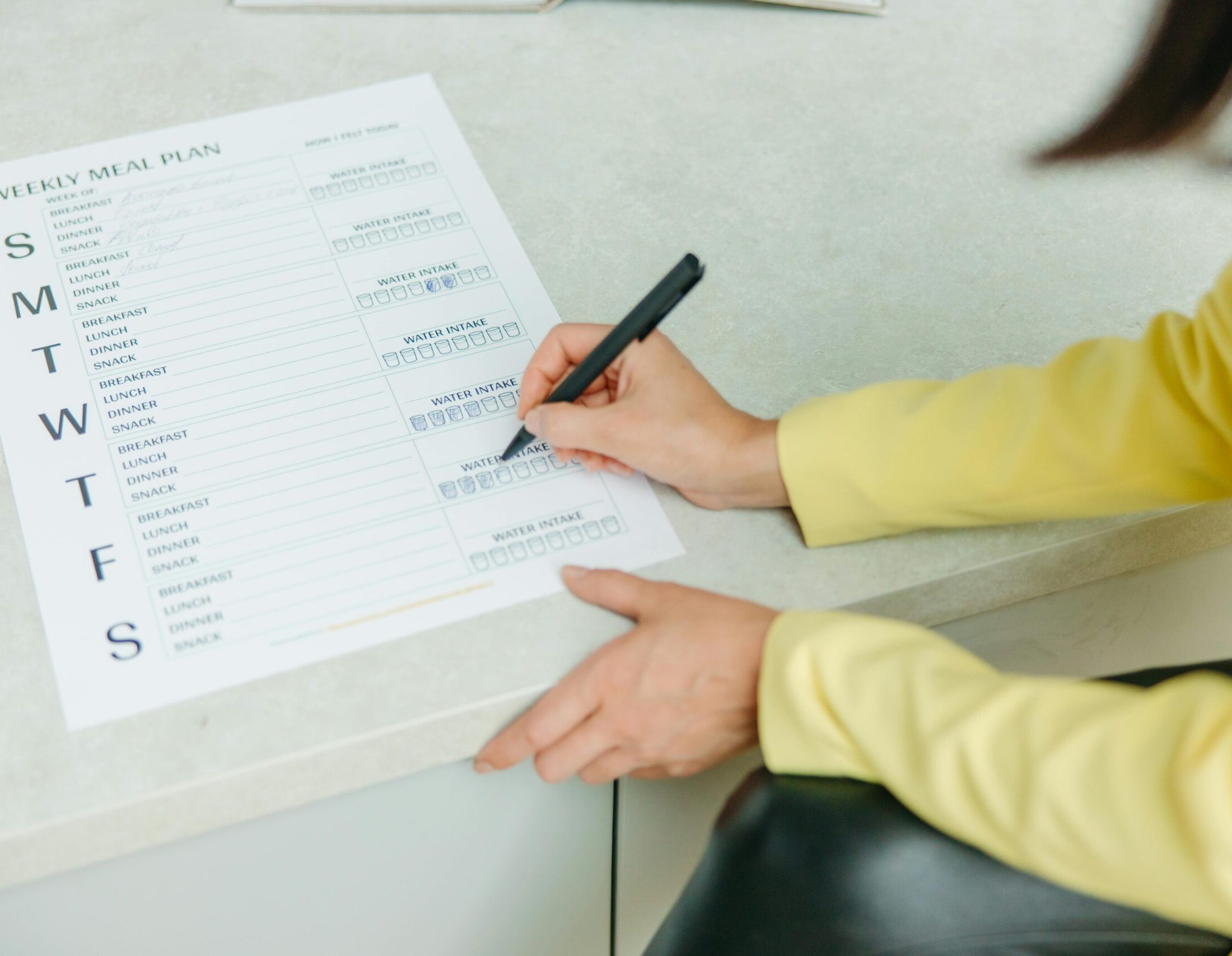 A close-up, over-the-shoulder view of a person wearing a vibrant yellow blazer filling out a printed 'Weekly Meal Plan' template with a black pen. The document, resting on a light stone countertop, includes sections for tracking daily breakfast, lunch, dinner, snacks, and water intake.
