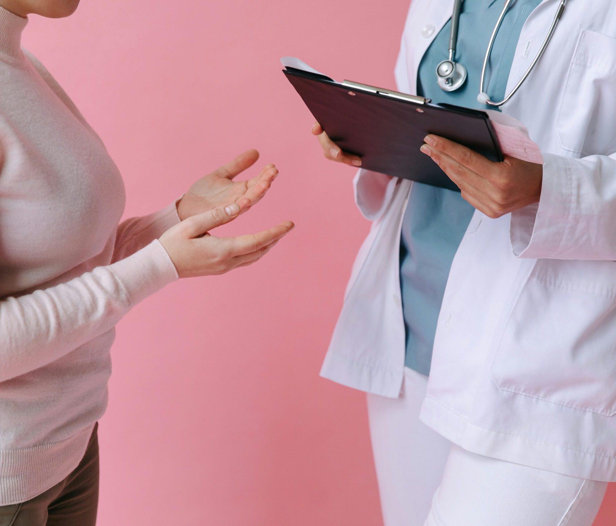 A close-up of a patient in a pink sweater gesturing with her hands while explaining her symptoms to a healthcare professional. The practitioner, wearing a white lab coat and stethoscope, holds a black clipboard and listens intently against a soft pink background, illustrating a patient-centered clinical consultation.